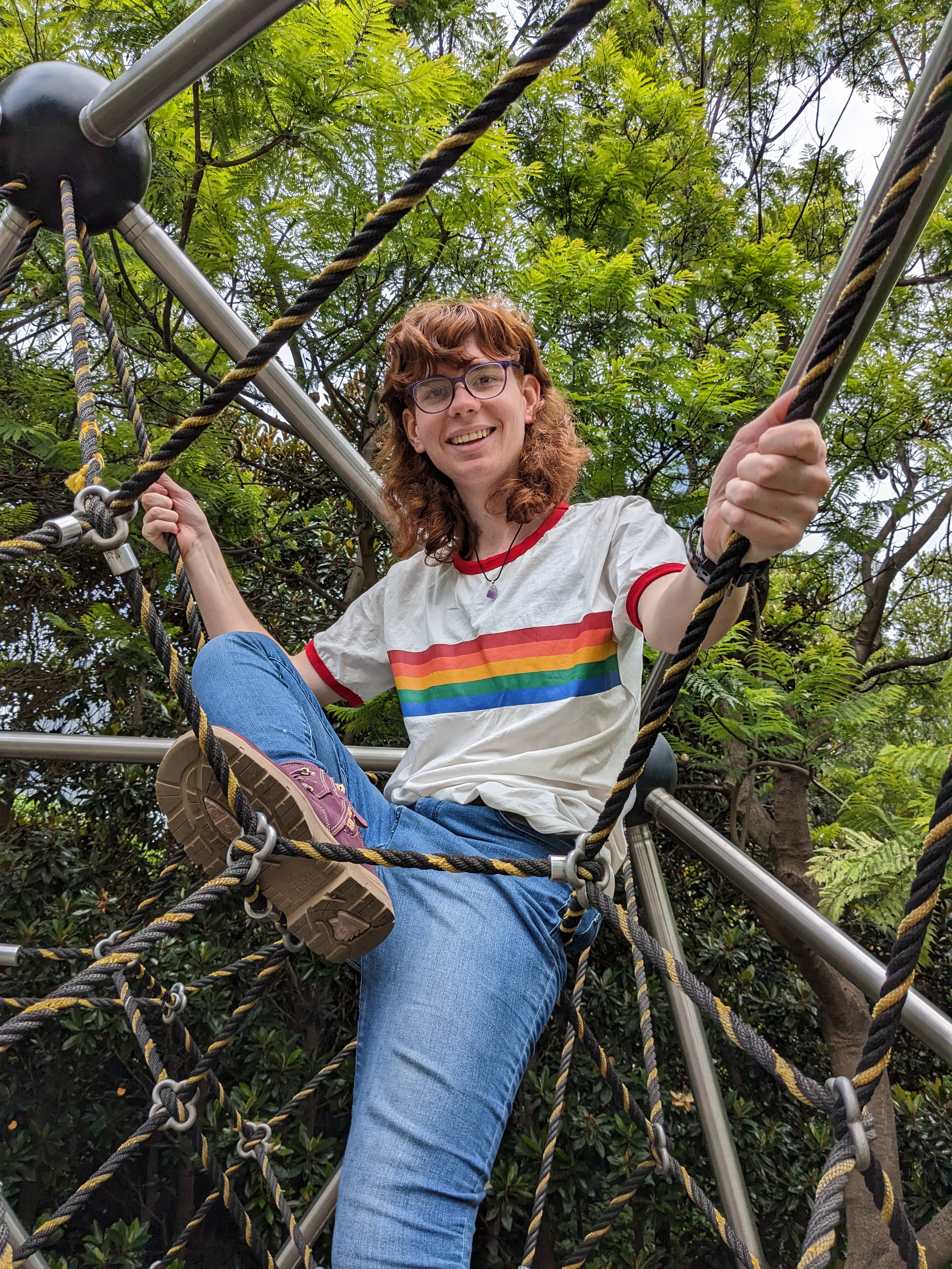 A young woman with red hair and a white shirt with a rainbow on it smiles down at the camera from on top of some playground equipment