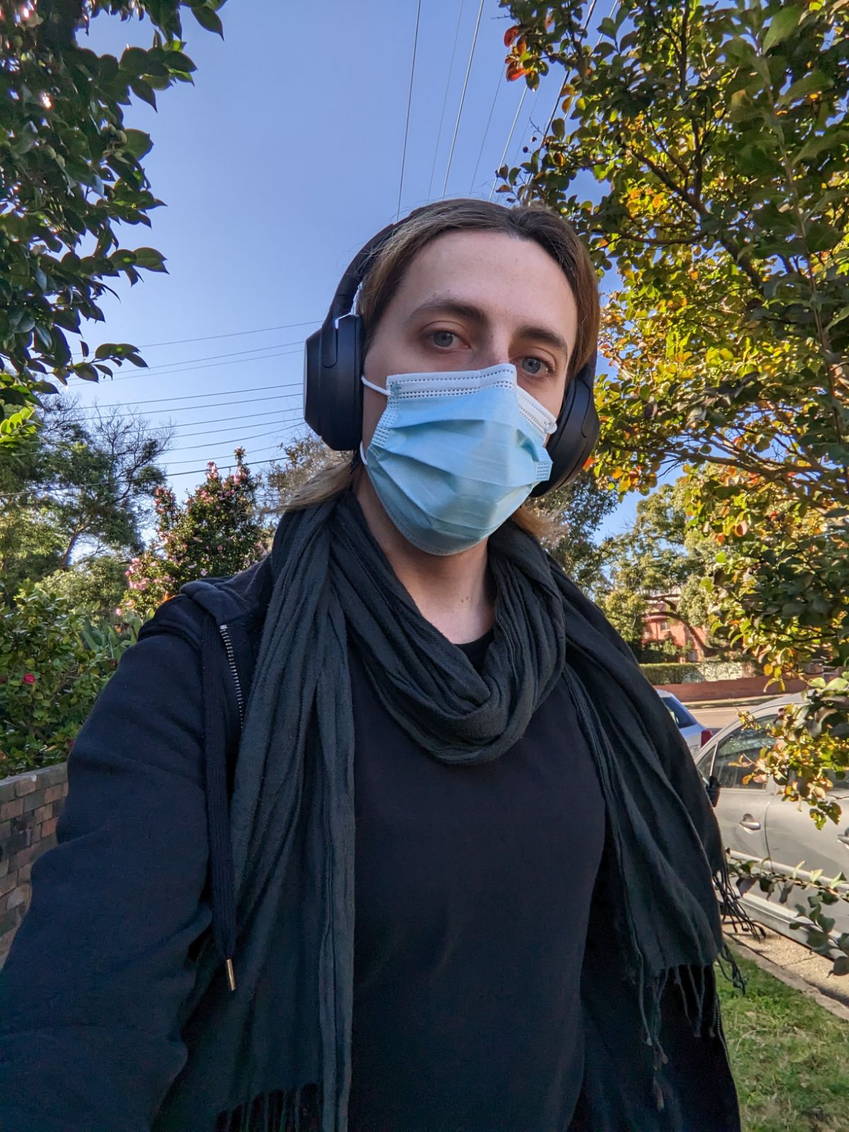 A woman is looking at the camera, wearing a mask, with trees and a blue sky in the background